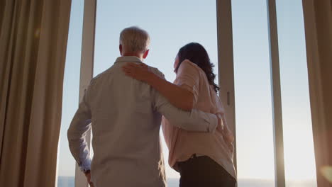 Glückliches-Paar,-Das-Sich-Im-Hotelzimmer-Umarmt-Und-Bei-Sonnenuntergang-Aus-Dem-Fenster-Blickt-Und-Im-Urlaub-Einen-Erfolgreichen-Ruhestandslebensstil-Genießt-Und-Eine-Romantische-Verbindung-Teilt