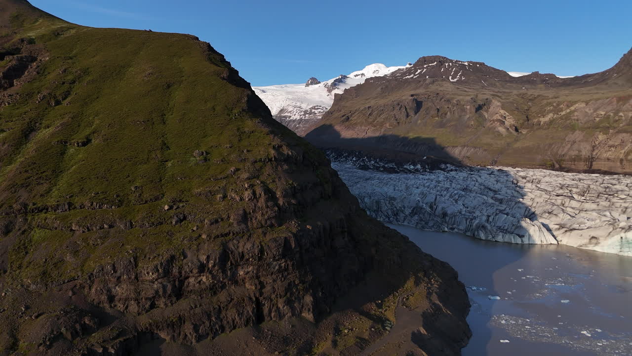 Aerial view of Svínafellsjökull Glacier at sunset, showing deep ice crevasses flowing into a dark glacial lagoon, framed by steep volcanic mountains and warm evening light in southern Iceland