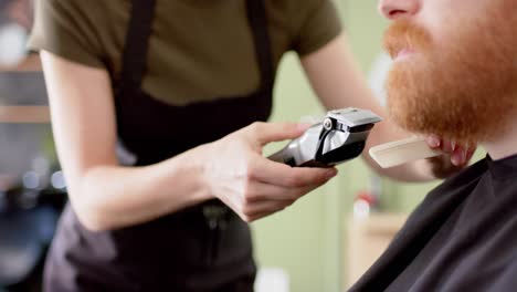 midsection of caucasian female barber trimming beard of male client at barbershop, in slow motion