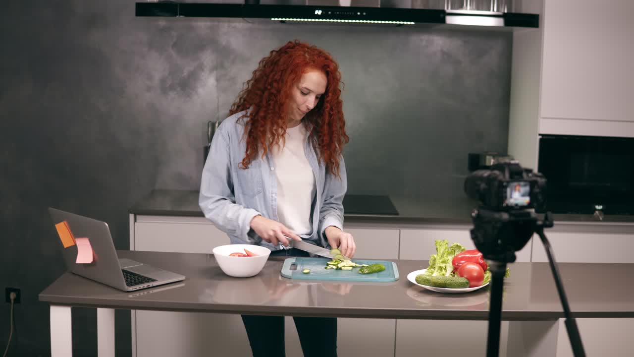 Young woman in casual clothes leads a culinary blog and records video in grey kitchen's bar. Video shooting of the blog - red headed cutting tomatos on a dashboard and explaining a recipe. Modern marketing, social networks