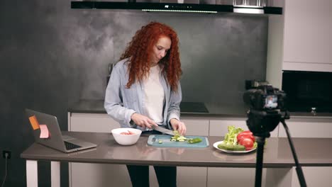 Young woman in casual clothes leads a culinary blog and records video in grey kitchen&#039;s bar. Video shooting of the blog - red headed cutting tomatos on a dashboard and explaining a recipe. Modern marketing, social networks