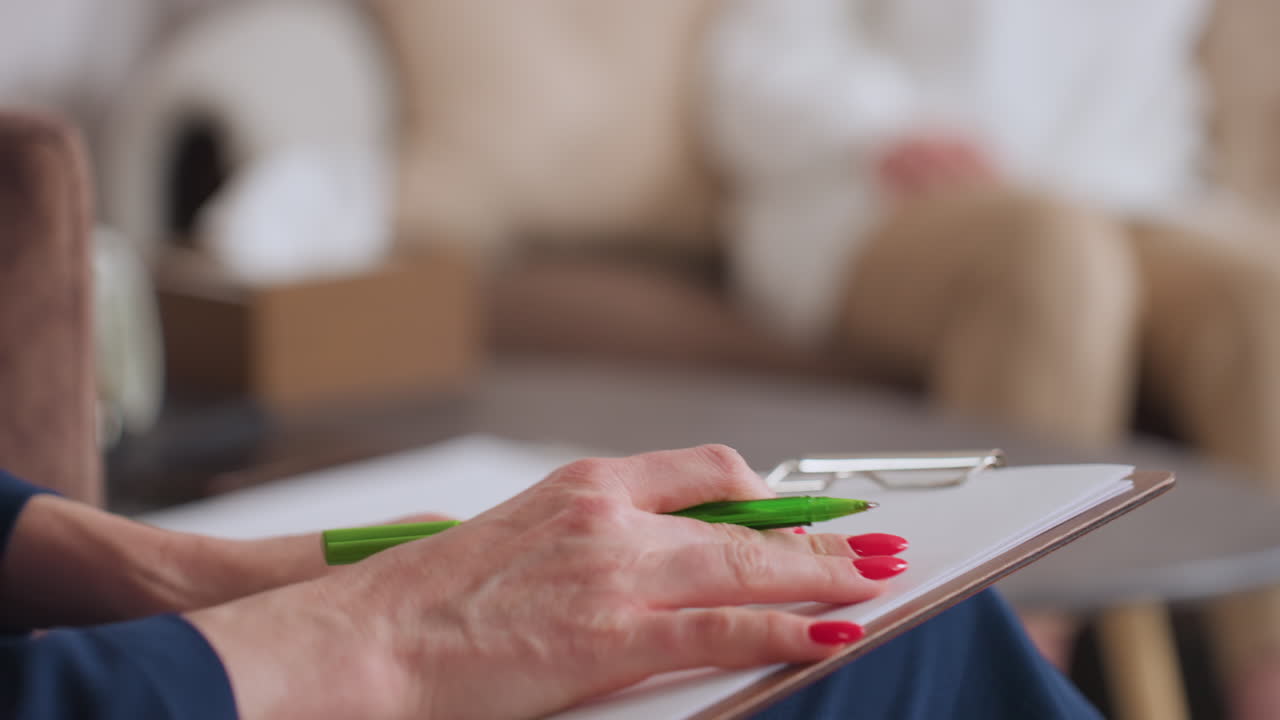 Close up hand view of counselor seated calmly writing on clipboard with green pen during therapy session, showing red manicured nails and gentle grip, blurred background includes soft beige cushions