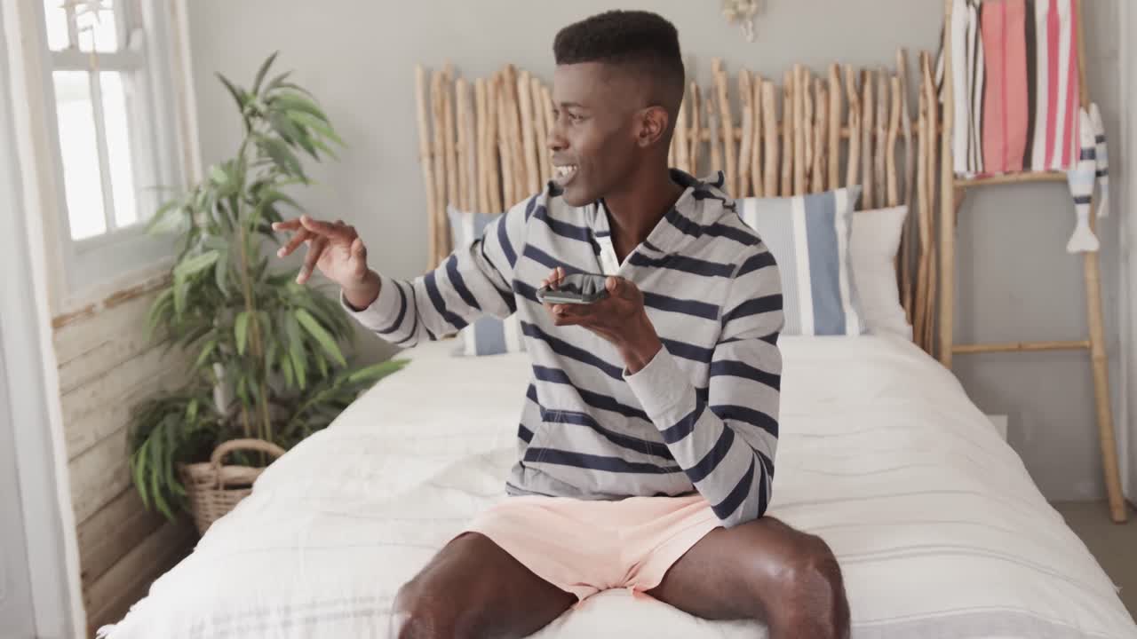 Happy african american man wearing shorts sitting on bed talking on smartphone, slow motion