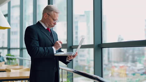 mature senior serious businessman with laptop in hands thinking while checking e-mails standing near window with panoramic city view. video from the side.