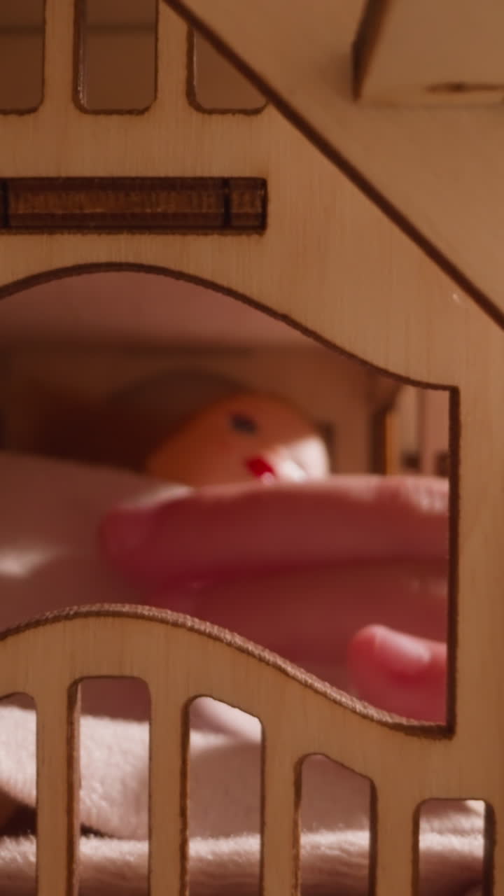 Little child hand taps doll lying under blanket on bed in wooden toy house with stairs and arch window on dark background close view