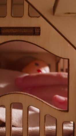 little child hand taps doll lying under blanket on bed in wooden toy house with stairs and arch window on dark background close view