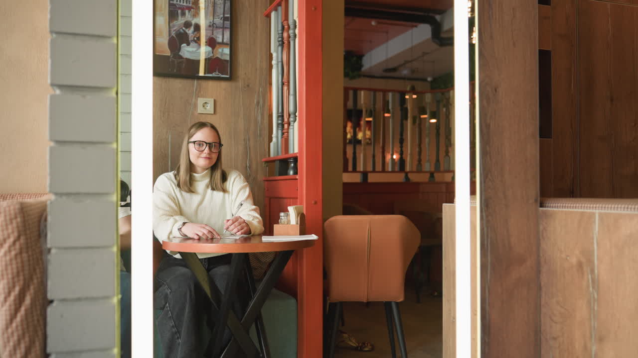 Female artist in white sweater and glasses sketching on paper at cafe table with soft indoor lighting, surrounded by wood and warm tones, focused on artwork while seated