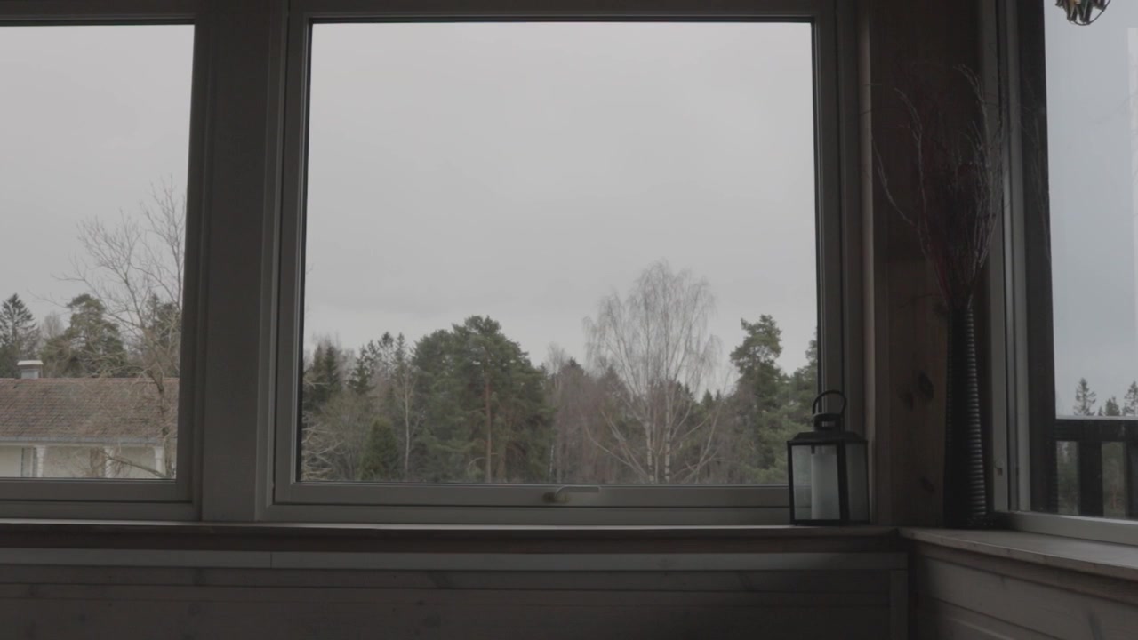 Interior view looking out through large window panes with a black lantern on the sill, revealing the moody, snow-dusted trees and overcast sky of Ekebergparken sculpture park in Oslo, Norway.
