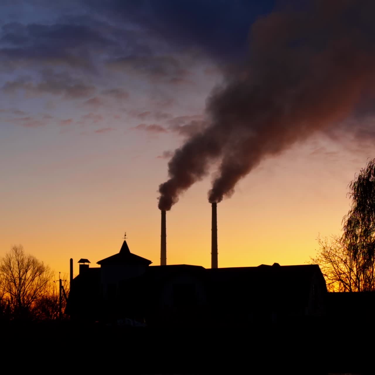 Beautiful autumn sky at sunset with silhouettes of a building, trees and plant pipes. Dark smoke goes up from the pipes