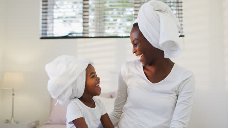 african american mother and daughter wearing towels sitting on bed and laughing