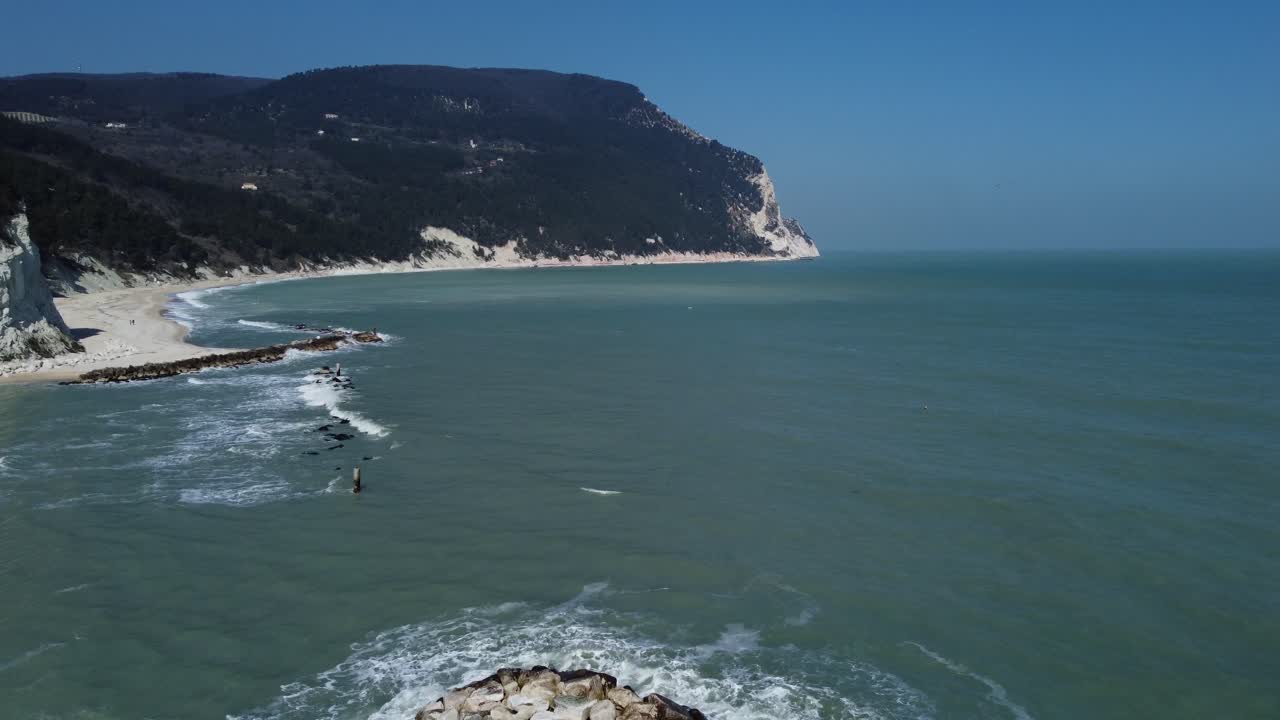 Drone flight up over sand beach with rocks, cliffs, green forrest hills and Adriatic sea waves, clear sky, early spring, a couple walks on the beach.