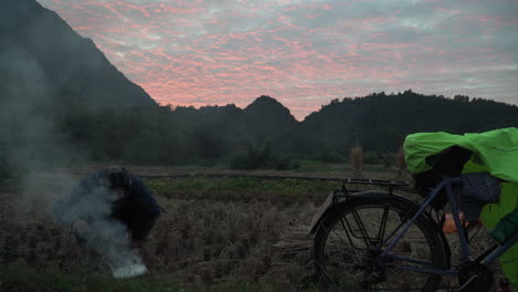 giovane uomo asiatico che fa un falò in campagna al tramonto con una bicicletta