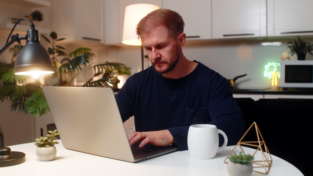 Giovane freelance apre il portatile di sera, inizia a lavorare da remoto, beve tè caldo, concentrato, calmo, a tarda notte