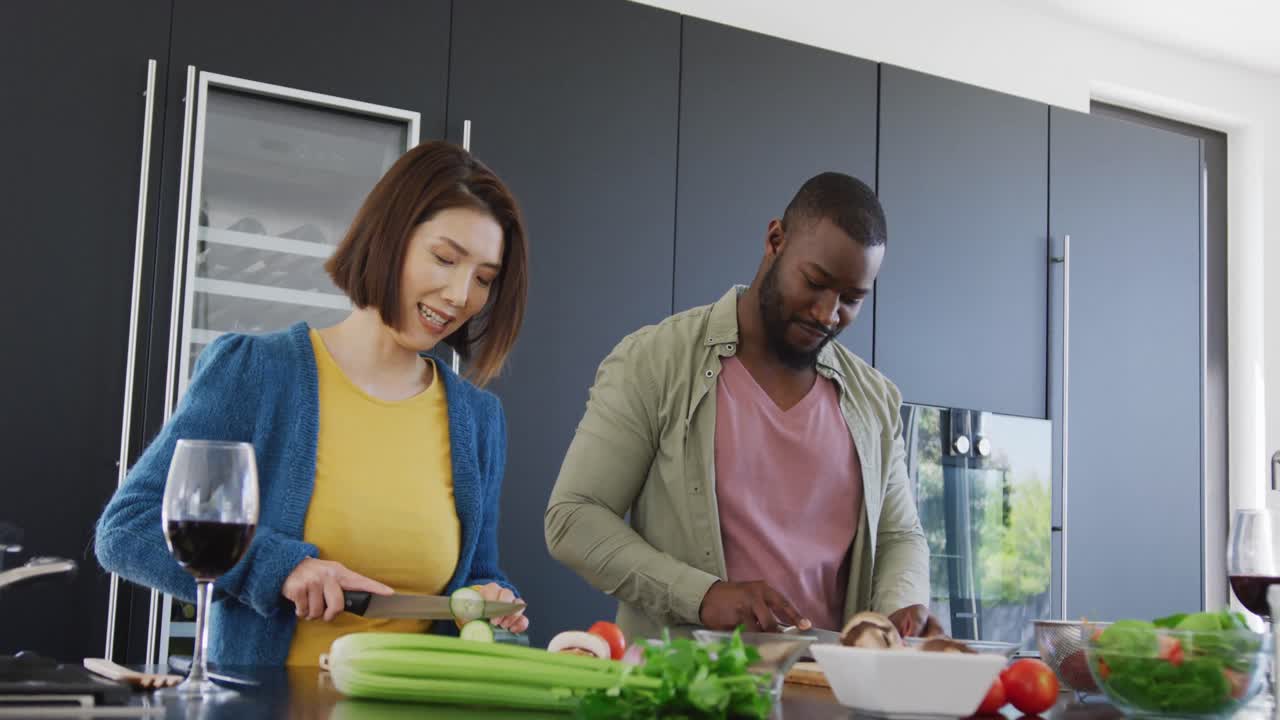 video de una feliz pareja diversa preparando comida y sonriéndose el uno al otro en la cocina en casa