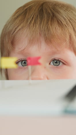 toddler boy looks at pins with color flags in notebook hiding behind table closeup. little child studies bright tag markers in copybook at lesson