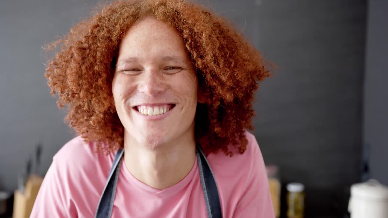 retrato de un hombre feliz de dos razas con el cabello rizado rojo usando un delantal sonriendo en la cocina, en cámara lenta.