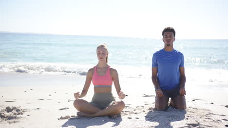 young caucasian woman and biracial man meditate on a sunny beach