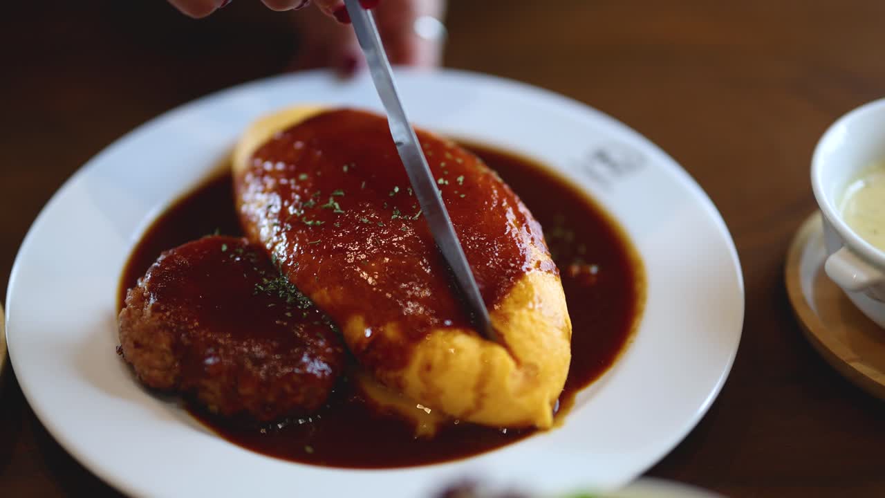 Hand slices omurice with demi-glace sauce beside hamburg steak, natural lighting, close-up view