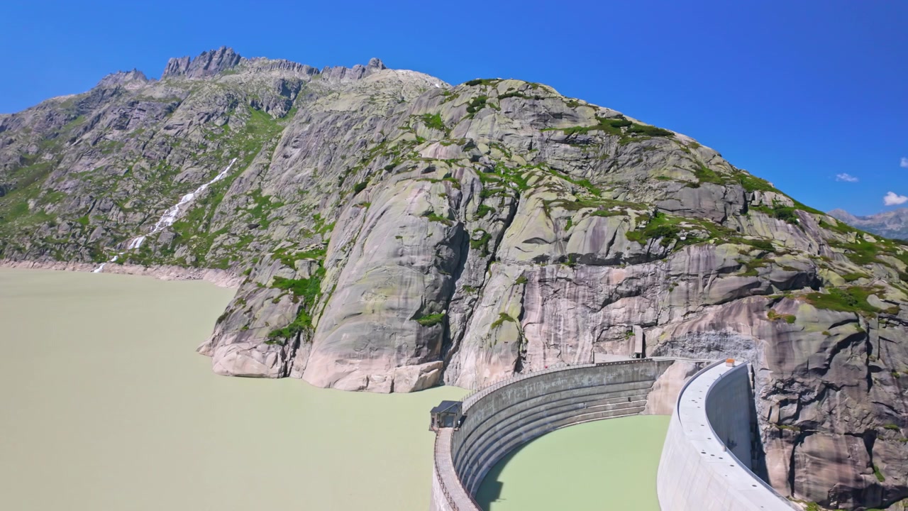 Bergsreservoar vid Grimselpasset med en böjd betongdamm, blekgrönt vatten och branta klippor under en klarblå himmel, som visar alpint landskap, ingenjörskonst och avlägsna sommarresor