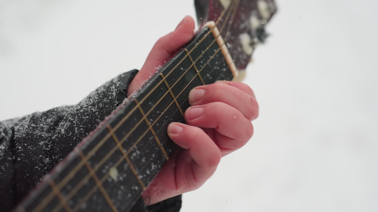 눈 인 현악기 위에서 기타를 연주하는 손의 클로즈업, 음악과 겨울의 평온한 아름다움 사이의 완벽한 조화를 만들어, 검은색 수갑과 기타 목에 얼어붙은 텍스처와 함께.