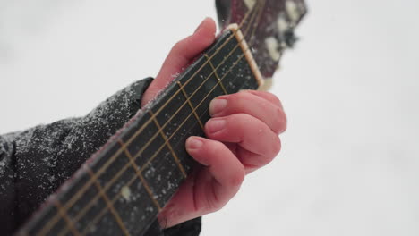 primer plano de la mano tocando la guitarra en cuerdas cubiertas de nieve, creando una armonía perfecta entre la música y la belleza serena del invierno, con texturas heladas en la manga negra y el cuello de la guitarra