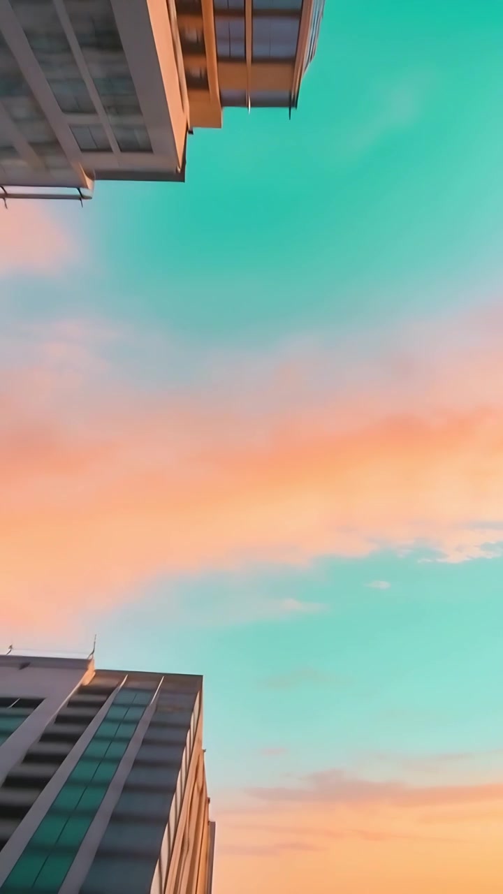 Vídeo vertical: Celular inclinando capturando nuvens do nascer do sol preenchendo céu turquesa entre arranha-céus.