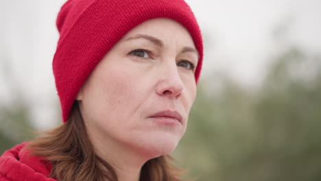 close-up of woman in red beanie and hoodie gazing thoughtfully into distance with serene expression, set in misty outdoor atmosphere