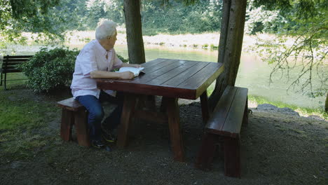 older man reads book at wooden table under trees near river