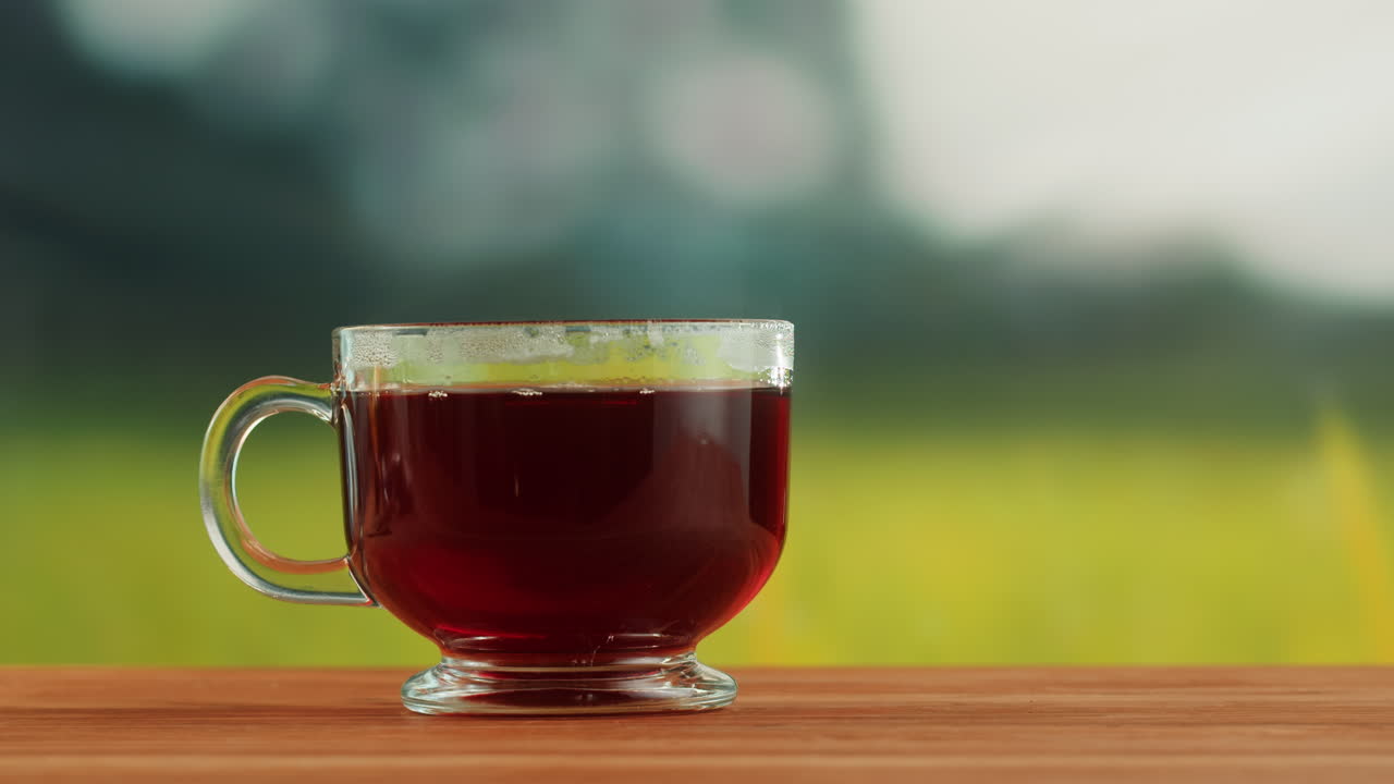 Steaming hot tea in a glass cup on a wooden table with a blurred natural background