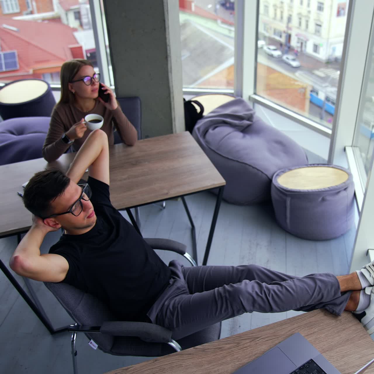 Relax from work in the office. Man stretched in his chair, his legs on the table. Female colleague drinking coffee and speaking on the phone. High angle view. City view in the window