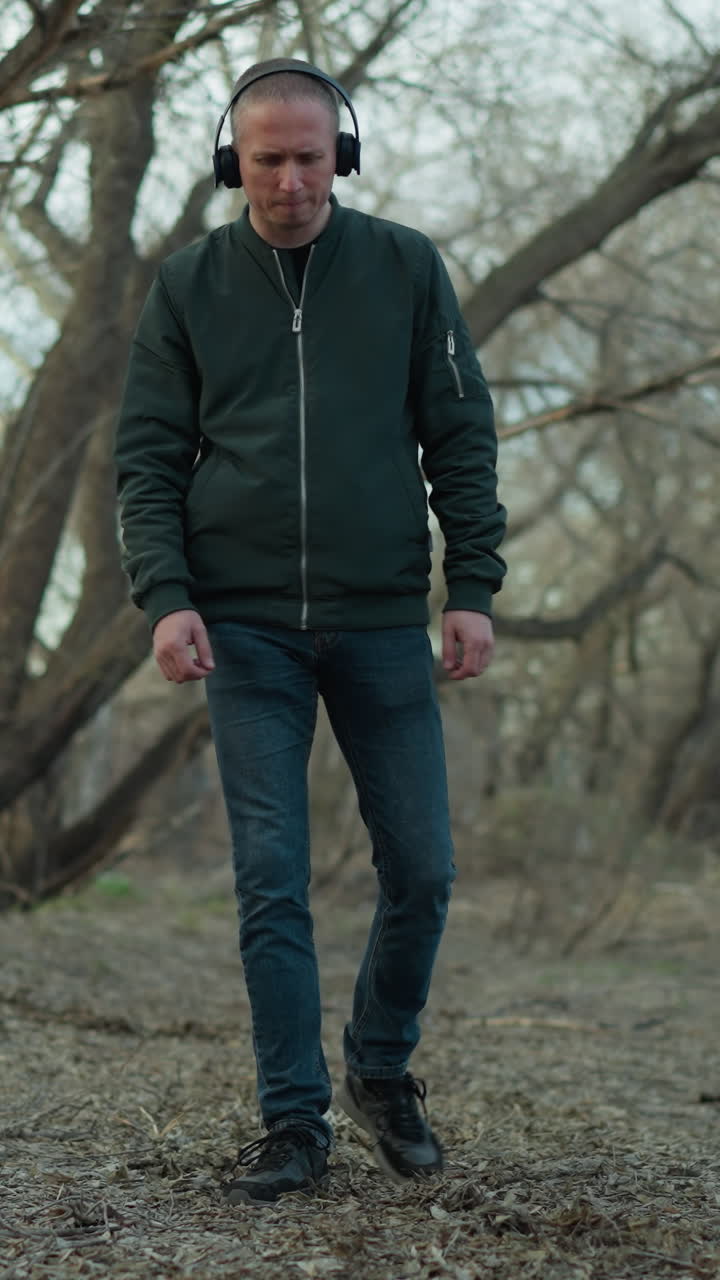 Man wearing a green jacket and headphones walking alone in a serene forest path, with the man seemingly lost in thought or enjoying his music as he strolls through the leaf-covered ground