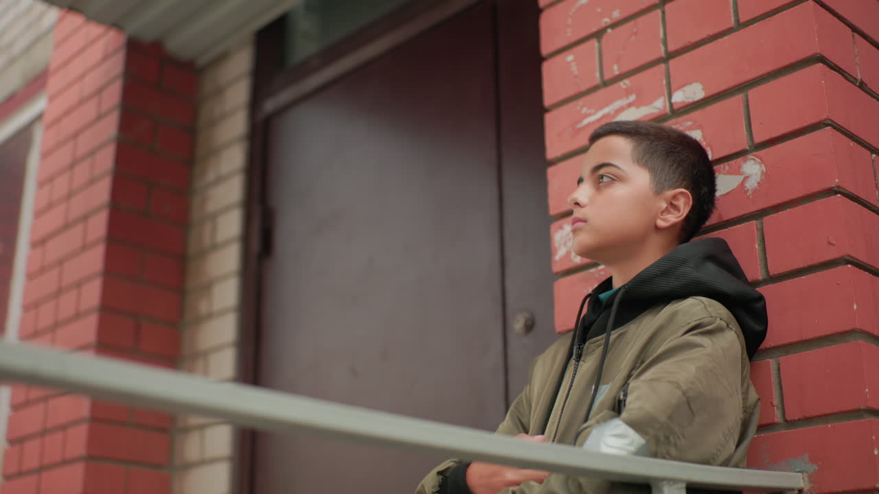 Calm kid in hooded jacket leans on red brick wall near doorway, turning head thoughtfully with focused expression, capturing quiet reflective mood of solitude outdoors