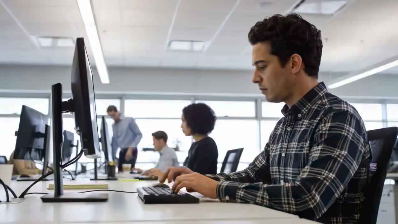 Typing man in plaid shirt glancing at monitor in office as colleague walking past, copy space