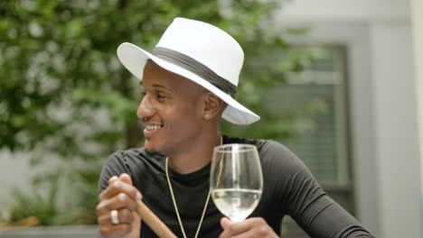 happy wealthy man wearing fedora hat and gold jewelry sits at a table holding wine holding a cigar outside near green plants socializes with others