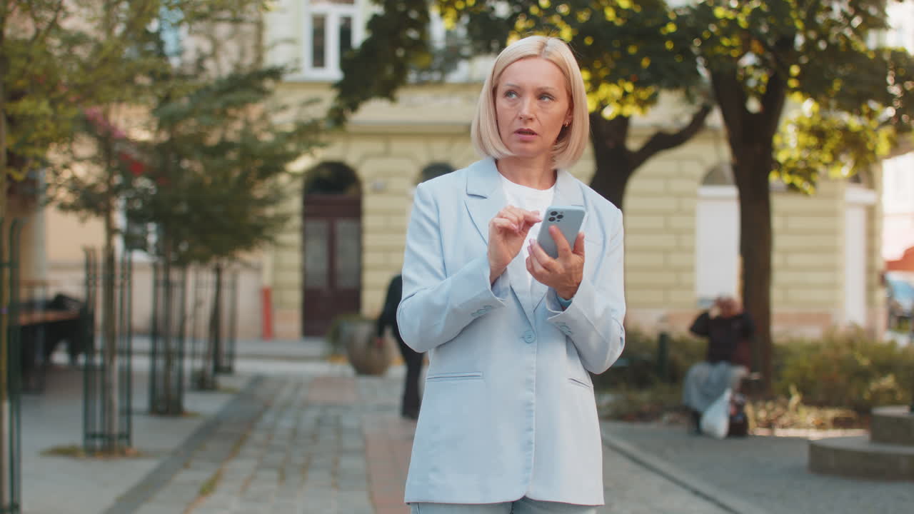 кавказская зрелая бизнесменка в костюме использует мобильный gps, чтобы найти направление, идущая по городской улице.