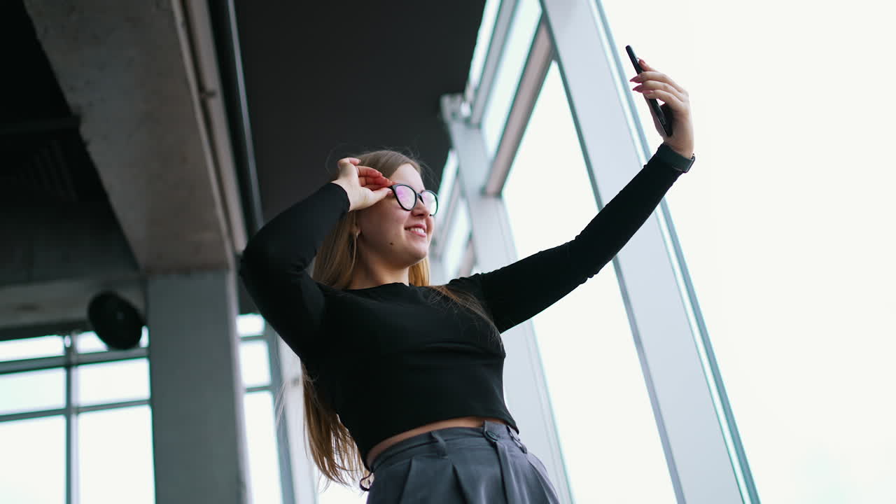 Attractive young woman in stylish glasses with a phone. Beautiful girl holding smart phone doing a selfie photo and smiles showing tongue. Panoramic windows background in office.