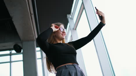 attractive young woman in stylish glasses with a phone. beautiful girl holding smart phone doing a selfie photo and smiles showing tongue. panoramic windows background in office.