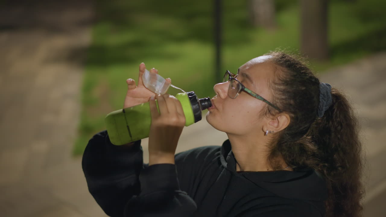 Young Woman Drinking Water Bottle Night Park Pathway Closeup Wearing Glasses And Hoodie, Reusable Green Bottle Sip Between Breaths, Cooling Down After Jog, Focused Calm Mood, Soft Bokeh Lights Urban