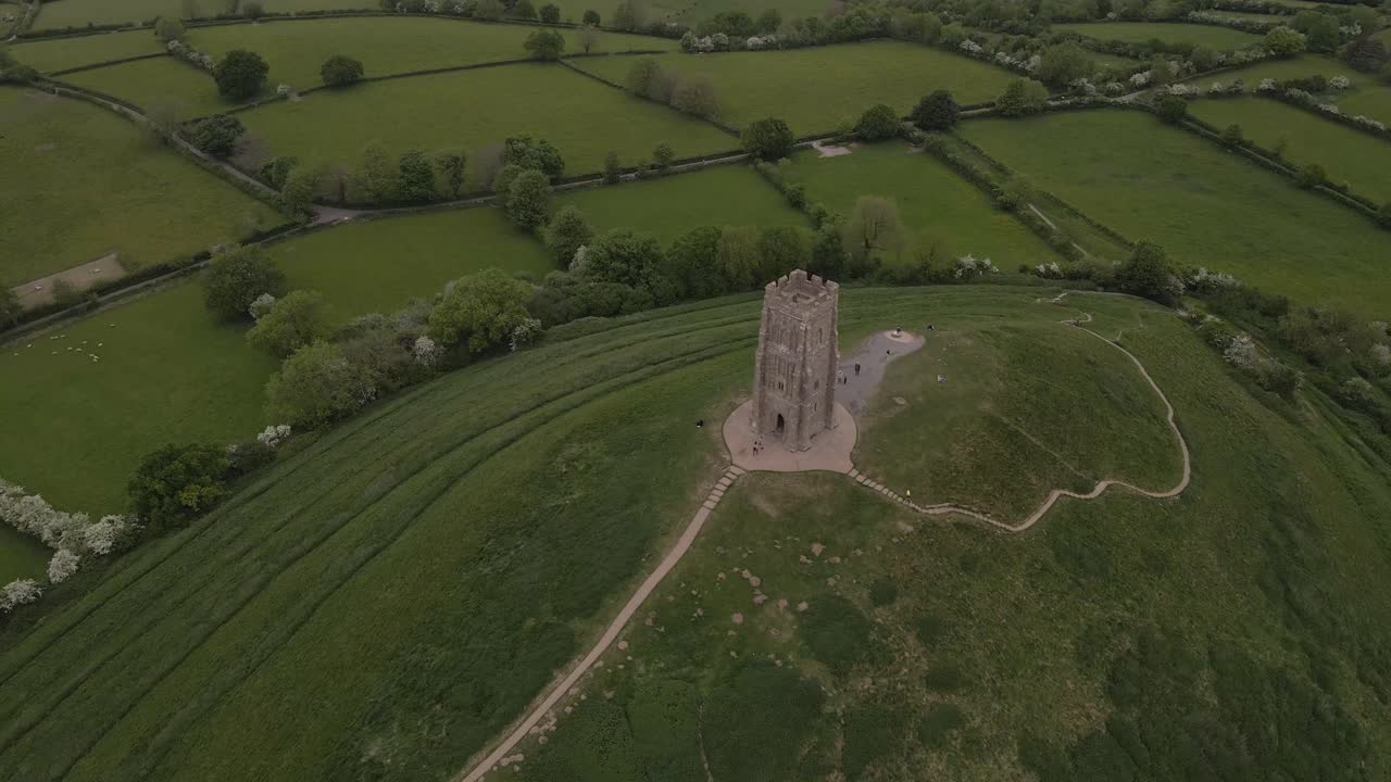 Aerial view of Glastonbury, drone moving in direction to the St Michaels tower and camera tilting down, drone flying over the green fields that surrounds the Tor. 4K, 60fps.