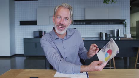 Smiling-caucasian-businessman-having-video-chat-going-through-paperwork-in-office-kitchen