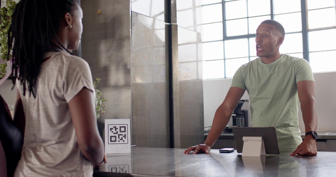 African American woman approaching counter, man tapping tablet processing payment, QR sign visible