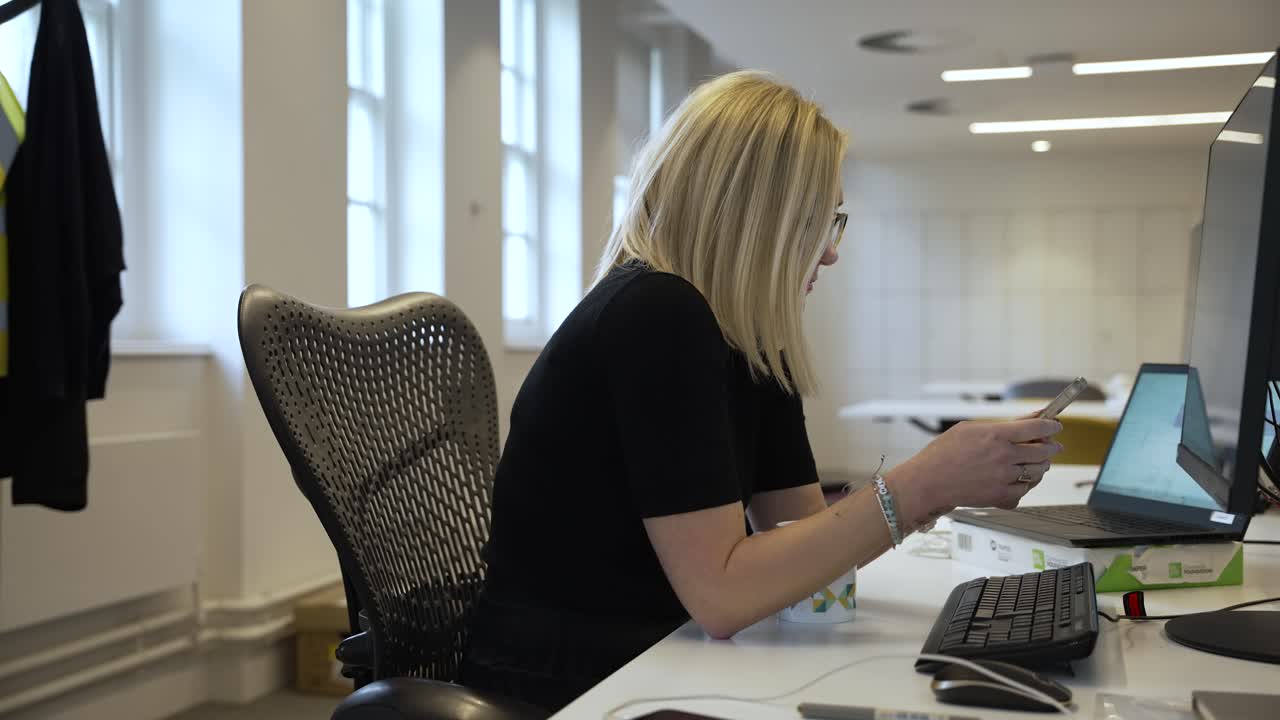 Female office worker sits in a professional office environment, browsing on a mobile phone while facing the computer monitor screen, modern multitasking and digital connectivity in the workplace