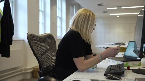 Female-office-worker-sits-in-a-professional-office-environment,-browsing-on-a-mobile-phone-while-facing-the-computer-monitor-screen,-modern-multitasking-and-digital-connectivity-in-the-workplace