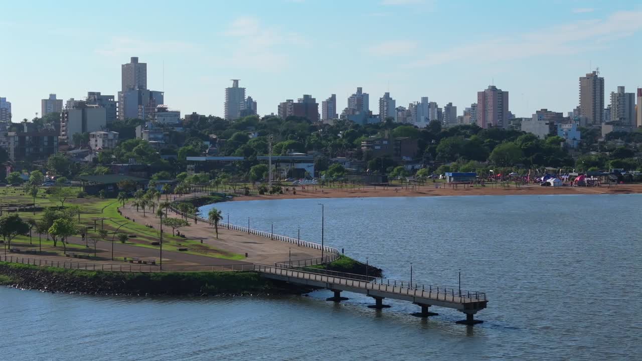 Close-up panning aerial captures sandy shoreline, green park areas, and waterfront infrastructure of Balneario El Brete in Posadas, Argentina, blending natural beauty with urban recreational design