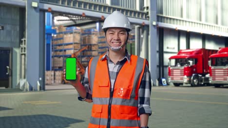 asian male engineer with safety helmet smiling and showing green screen smartphone to the camera while standing , outside of logistics distributions warehouse