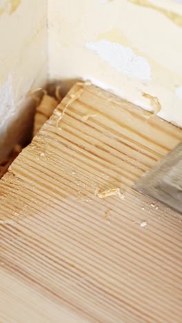 Person's hand using a metal scraper to remove old paint and filler from the corner of a wooden floor, revealing the natural grain and prepping surface for refinishing, extreme close-up vertical shot