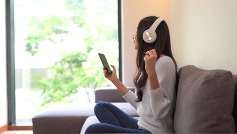 happy asian woman relaxing and using overhead headphones to listen to music from smart phone sitting on sofa and snaps fingers to the beat - side view