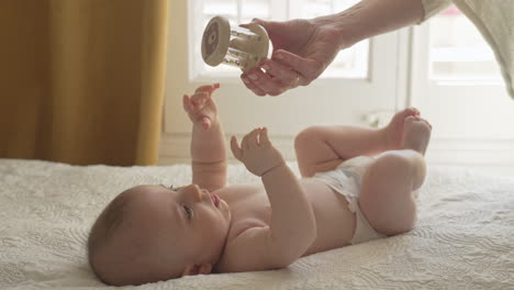 cute baby laying on his back in white bed sheets, looking at toy offered from mother, in bright bedroom