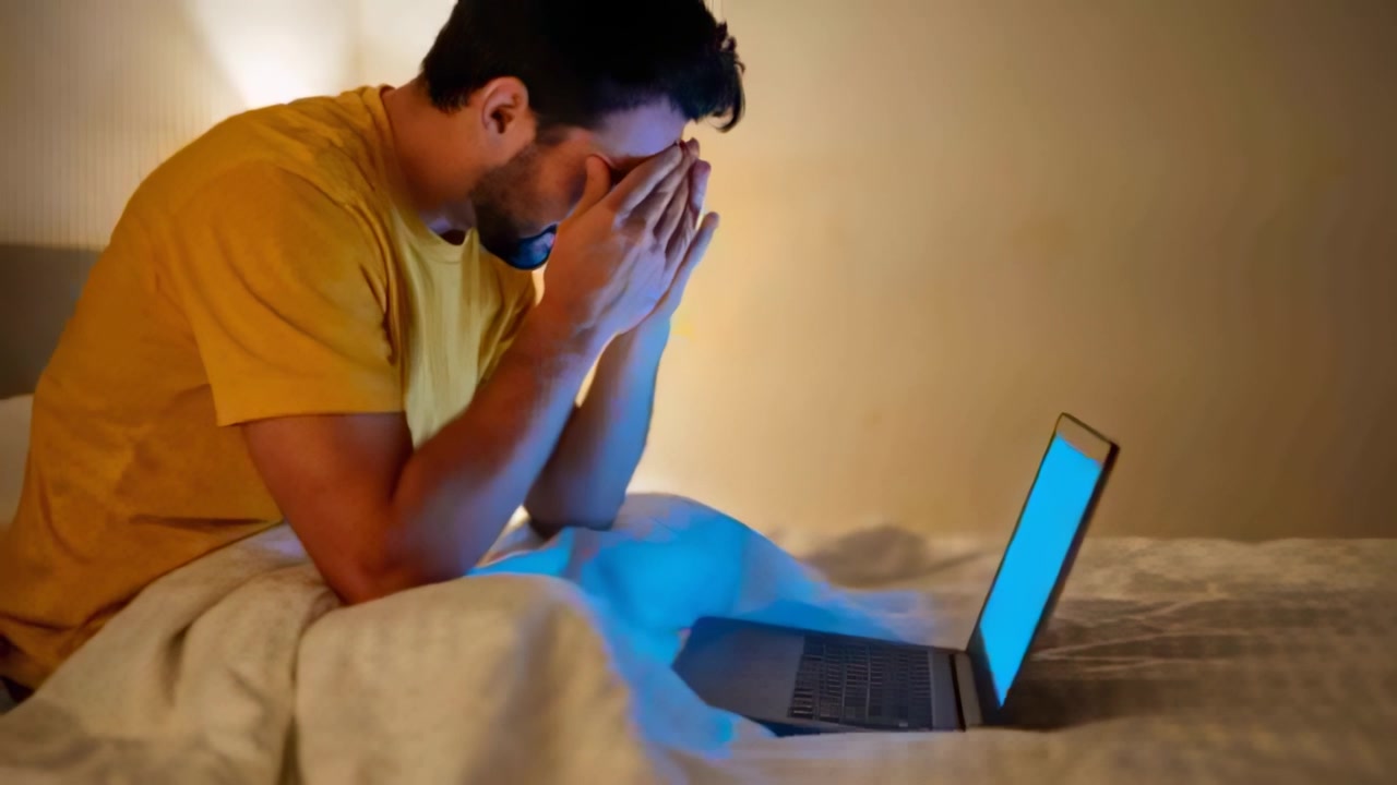 Reacting to bright laptop screen, man rubbing face on bed, seeking relief, wearing yellow tee