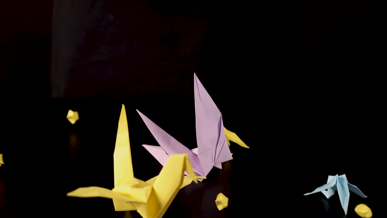 Yellow and pink origami cranes drop small paper stars, dramatic lighting, static camera, black background